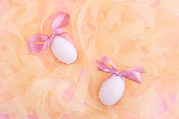 ピンクのかわいい弓を持つ2つの白い卵は 光沢のあるリボンのヒープに横たわっています ミニマルでエレガントなイースターフラットレイデザイン ストックフォト