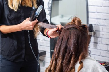 Modern bir kuaförde müşterilerin saçlarını kıvırmak için ütü kullanan profesyonel bir kuaför. Kuaför, saçını kestirirken beyaz kadın için özel muamele uyguluyor.