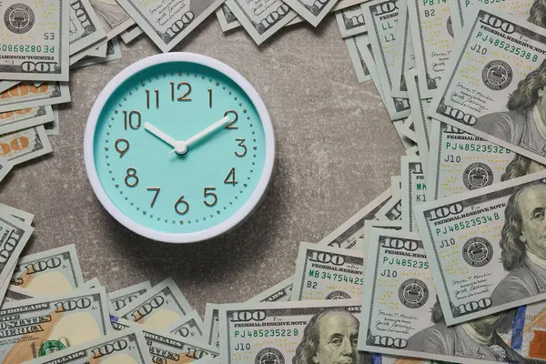 Round analog clock and a lot of hundred dollar bills on a gray background. Top view.
