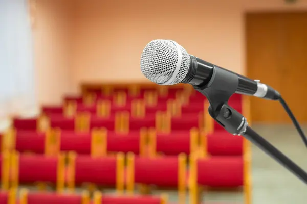 A microphone on a stand and an empty auditorium in a blurred background. Indoors.