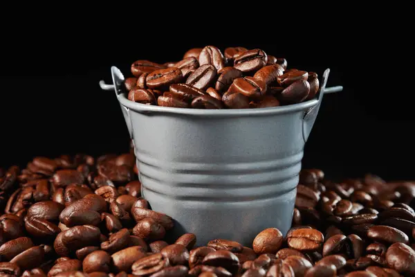 A bucket of roasted coffee beans in a pile of coffee beans on a black backdrop. Front view from low angle.
