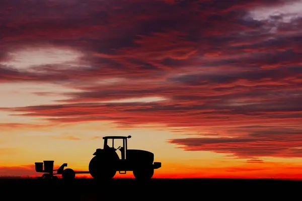 Silhouette of a tractor with a seeder against a bright sunset sky with dramatic clouds, depicting rural life and farming.