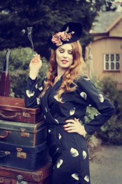 Eski moda elbiseli bir kadın tren istasyonunda eski tarz bavullarla poz veriyor.