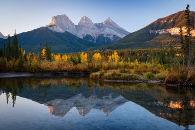 Sonbaharda şafak vakti Kanada Kayalıkları 'nın güzel manzarası. Dağ, sarı ve yeşil renkli orman suya yansıyor. Canmore, Alberta, Kanada. Üç Kız Kardeş Tepeler Üçlüsü.