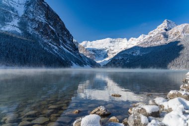 Louise Gölü kışın erken saatlerinde güneşli bir günde. Turkuaz renkli su yüzeyinde süzülen sis. Açık mavi gökyüzü, arka planda karla kaplı dağlar. Banff Ulusal Parkı 'nda güzel doğal manzara.