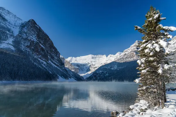 Louise Gölü kışın erken saatlerinde güneşli bir günde. Turkuaz renkli su yüzeyinde süzülen sis. Açık mavi gökyüzü, arka planda karla kaplı dağlar. Banff Ulusal Parkı 'nda güzel doğal manzara.
