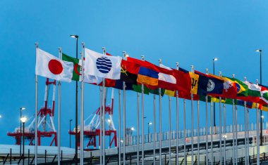 Uluslararası bayraklar, 2025 Osaka Kansai Fuarı 'nın doğu kapısı girişinde açık bir gökyüzüne karşı dalgalanıyor. Osaka, Japonya
