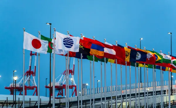 Uluslararası bayraklar, 2025 Osaka Kansai Fuarı 'nın doğu kapısı girişinde açık bir gökyüzüne karşı dalgalanıyor. Osaka, Japonya