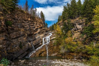 Cameron Falls güneşli bir sonbahar sabahında mavi gökyüzü ve dağların üzerinde beyaz bulutlarla. Waterton Lakes Ulusal Parkı, Alberta, Kanada.