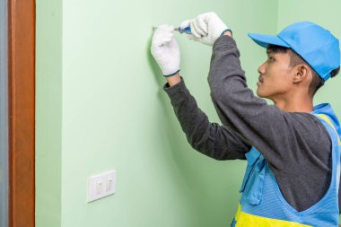 Asyalı erkek işçi alçı taşı duvarındaki vidaları sıkmak için tornavida kullanıyor.