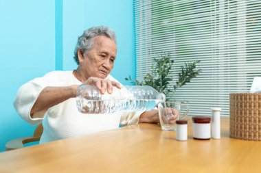 Yaşlı bir Asyalı kadın ilaç almaya hazırlanmak için plastik şişeden bardağa su dolduruyor..