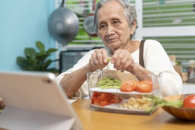 Yaşlı bir kadın mutfağında yemek pişiriyor ve yemek yapmayı öğrenmek için dijital tabletlere bakıyor..