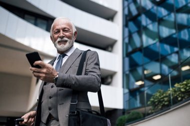 Akıllı telefon kullanarak gülümseyen kıdemli iş adamının portresi. İş seyahati sırasında dizüstü bilgisayar çantası taşıyan yaşlı profesyonel bir erkek takım elbise giyiyor. Şehirdeki ofis binasına karşı duruyor..