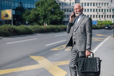 Üst düzey işadamı akıllı telefonda konuşurken dizüstü bilgisayar çantası taşıyor. Yaşlı profesyonel erkek, kentte yol kenarında bekliyor. Güneşli bir günde takım elbise giyiyor..