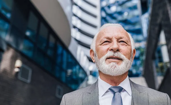 Close-up of thoughtful senior businessman with beard. Elderly contemplating male professional is standing against modern office building in city. He is wearing suit.