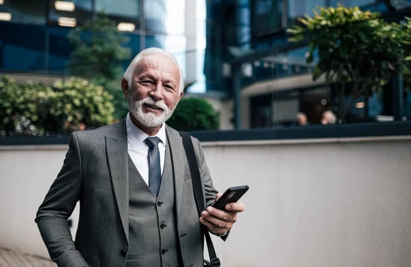 Portrait of smiling elderly businessman using smart phone. Elderly male professional wearing suit is carrying laptop bag in the city. He is standing against modern office building during travel.