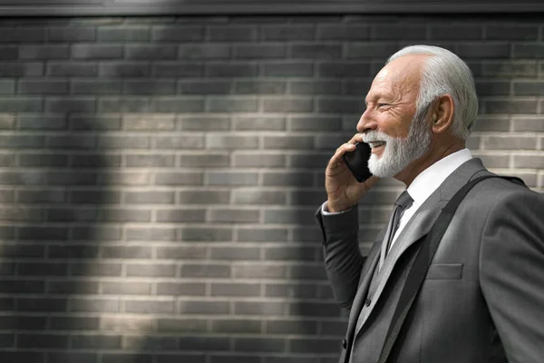 Profile view of smiling senior businessman talking on smart phone. Elderly male entrepreneur with bag standing by brick wall. He is wearing suit while traveling in the city.