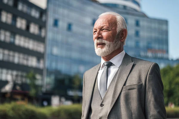Thoughtful senior businessman looking away. Serious elderly professional is standing against modern buildings in the city. He is wearing full suit during business travel on sunny day.