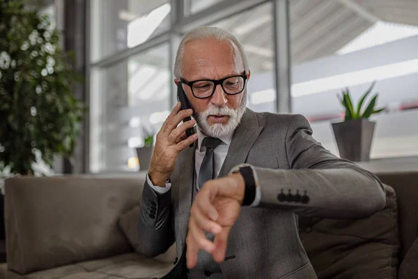 Elderly businessman checking time on wristwatch. Senior male professional wearing eyeglasses is talking on smart phone. He is sitting on sofa at corporate office.