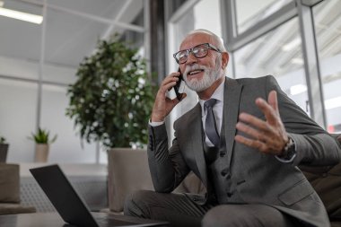 Akıllı telefondan konuşurken neşeli kıdemli profesyonel jestler. Yaşlı girişimci kanepede otururken masada dizüstü bilgisayar kullanıyor. İş yerinde takım elbise giyiyor..