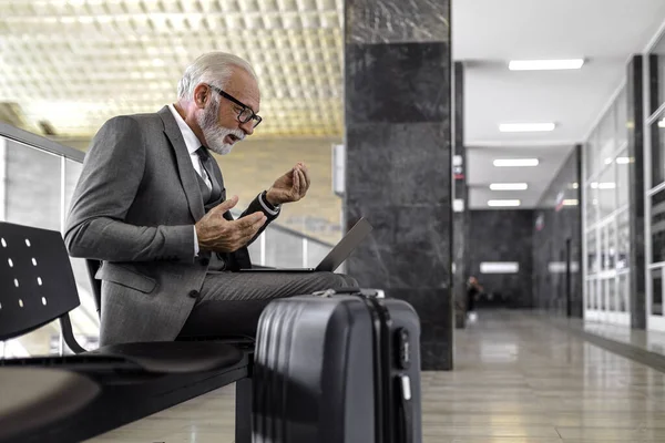 Confident elderly businessman gesturing while talking through video call on laptop. Senior professional is sitting on chair while waiting at the train or subway station. He is wearing suit.