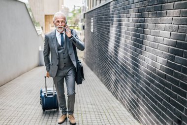 Kıdemli işadamı akıllı telefondan konuşuyor. Form giyen yaşlı erkek profesyonel dizüstü bilgisayar çantası taşıyor. Şehirdeki ofis binasına karşı yaya olarak yürüyor..