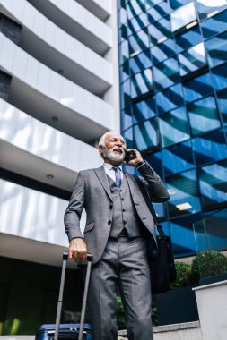 Zeki telefondan konuşan kıdemli bir profesyonelin düşük açılı görüntüsü. Takım elbiseli bir işadamı dizüstü bilgisayar çantası taşıyor ve bavulu tutuyor. Şehirdeki ofis binasına karşı duruyor..