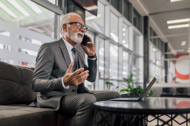 Akıllı telefonda konuşurken gülümseyen profesyonel erkek jestler. Kıdemli girişimci masada otururken dizüstü bilgisayar kullanıyor. Ofiste çalışırken takım elbise giyiyor..