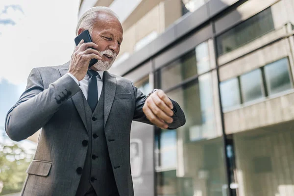 Kıdemli işadamı kol saatini kontrol ediyor. Erkek profesyonel takım elbiseli akıllı telefonda konuşuyor. Şirket binasına karşı duruyor..