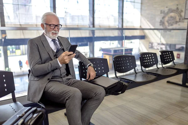 Smiling businessman with luggage socializing on smart phone. Senior male professional is sitting on chair while waiting at train or subway station. He is wearing suit during business travel.