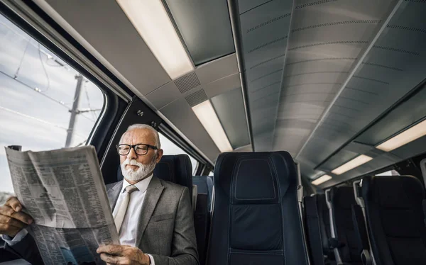 Senior entrepreneur reading newspaper while sitting by the window. Male professional is traveling in modern passenger train. He is wearing suit while commuting.