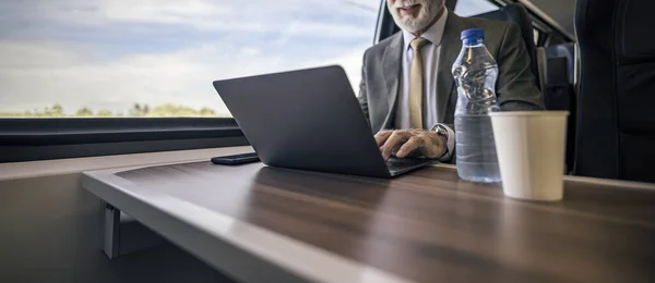 Midsection of elderly professional working on laptop. Bottle and disposable cup on table on passenger train. He is commuting through rail and train transportation.