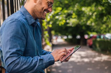 Neşeli yetişkin sakallı bir adamın tabletinde internette gezinen, dışarıda güzel bir günün tadını çıkaran bir resmi..