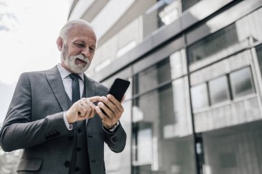 Akıllı telefonda gülümseyen profesyonel iş mesajları. Yaşlı girişimci ofis binasının önünde duruyor. Takım elbise giyiyor..