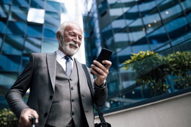 Gülümseyen kıdemli girişimci akıllı telefonda sosyalleşiyor. Takım elbiseli yaşlı işadamı dizüstü bilgisayar çantası taşıyor. İş seyahati sırasında modern ofis binasına karşı duruyor..