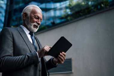 Dijital tabletle ilgili fikirleri analiz eden kendine güvenen bir iş adamı. Kablosuz bilgisayar kullanarak gülen kıdemli girişimci takım elbise giyiyor. Şehirdeki ofis binasına karşı duruyor..