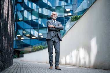 Üst düzey işadamları kollarını kavuşturup başka tarafa bakıyor. Düşünceli yaşlı profesyonel modern ofis binasının önünde duruyor. Takım elbise giymiş..