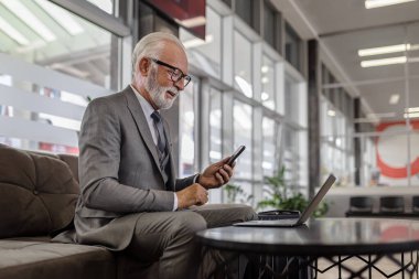 Otel binasında dizüstü bilgisayar kullanırken cep telefonuyla yaşlı profesyonel ağ kurma.