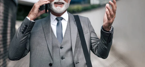 Midsection of happy professional talking on smart phone. Smiling businessman with bag gesturing while standing against office building. He is wearing full suit while commuting in the city.