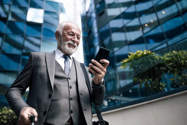 Gülümseyen kıdemli girişimci akıllı telefonda sosyalleşiyor. Takım elbiseli yaşlı işadamı dizüstü bilgisayar çantası taşıyor. İş seyahati sırasında modern ofis binasına karşı duruyor..