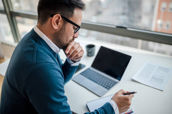 Thoughtful adult man, using a pen to write in the notebook, on his office desk.