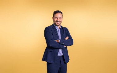 Yakışıklı, orta yaşlı, zarif mavi takım elbiseli, kolları çapraz gülümseyen ve turuncu arka planda poz veren işadamı.