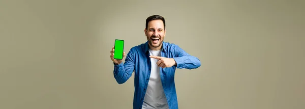 携帯電話の白いクロマキーの緑の画面を指し 灰色の背景に隔離された立っている間に陽気に笑って魅力的な中人男性 — ストック写真