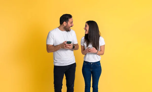 兴奋的年轻夫妇穿着白色T恤 面面相觑 一边欢呼雀跃 一边用黄色背景的智能手机庆祝胜利 — 图库照片