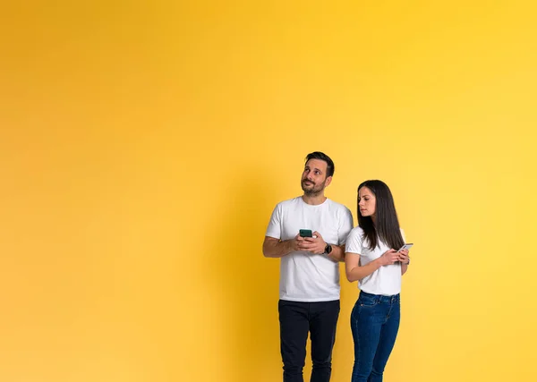 嫉妒的年轻成年女友一边站在黄色的背景下一边看着男朋友的智能手机一边聊天 — 图库照片