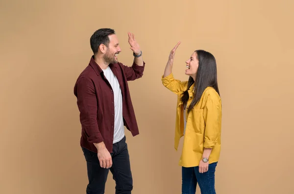 Ecstatic man and woman screaming and giving high five to each other while celebrating success. Excited young couple clapping hands and greeting while standing over yellow background