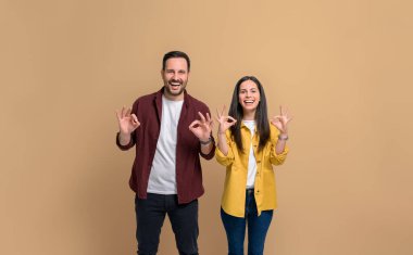 Gömlek giymiş genç bir kadın ve erkek bej arka planda poz verirken gülüyorlar ve iyi işaretler gösteriyorlar. Kameraya bakıp neşeli bir şekilde el kol hareketi yapan çiftin portresi.