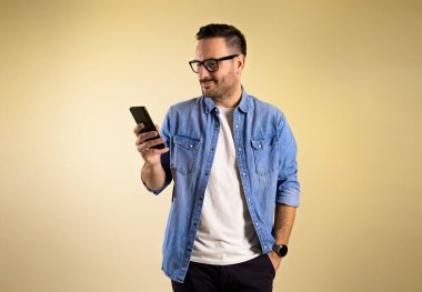 Bej arka planda tek başına dururken cep telefonunda mesajları kontrol eden çekici bir erkek serbest yazar. Gözlük ve kot pantolon giyiyor.