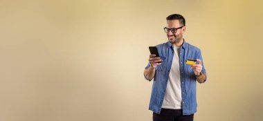 Akıllı telefon ve kredi kartıyla online alışveriş yapan gülümseyen erkek müşteri bej arka planda izole edilmiş. Genç adam e-ticaret uygulamasından alışveriş yapıyor ve mobil ödeme yapıyor.