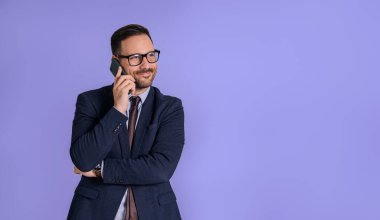 Zarif takım elbiseli genç işadamı telefonda konuşurken gülümsüyor ve yüzünü çeviriyor. Erkek profesyonel yönetici mavi arka planda izole akıllı telefondan konuşuyor.